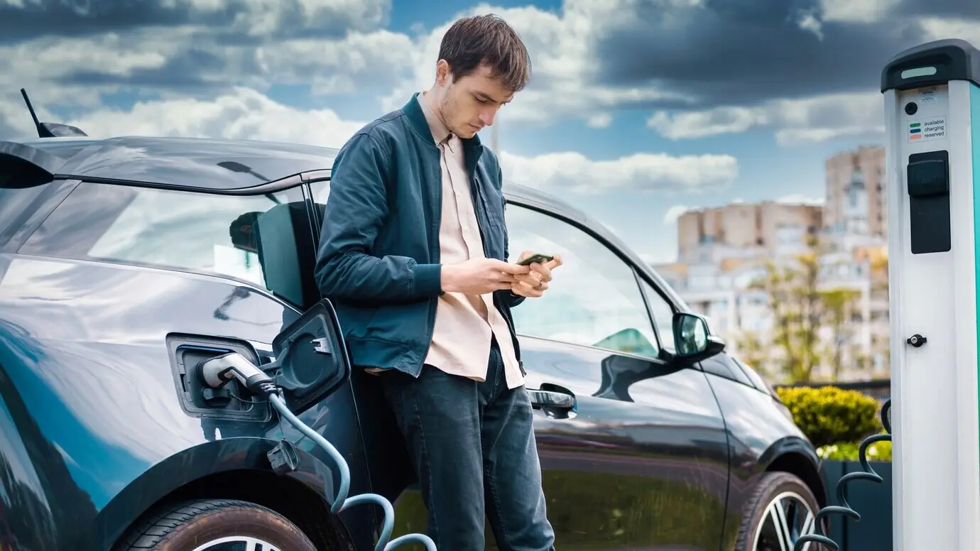 A man is charging his electric car at a charging station while using a smartphone.